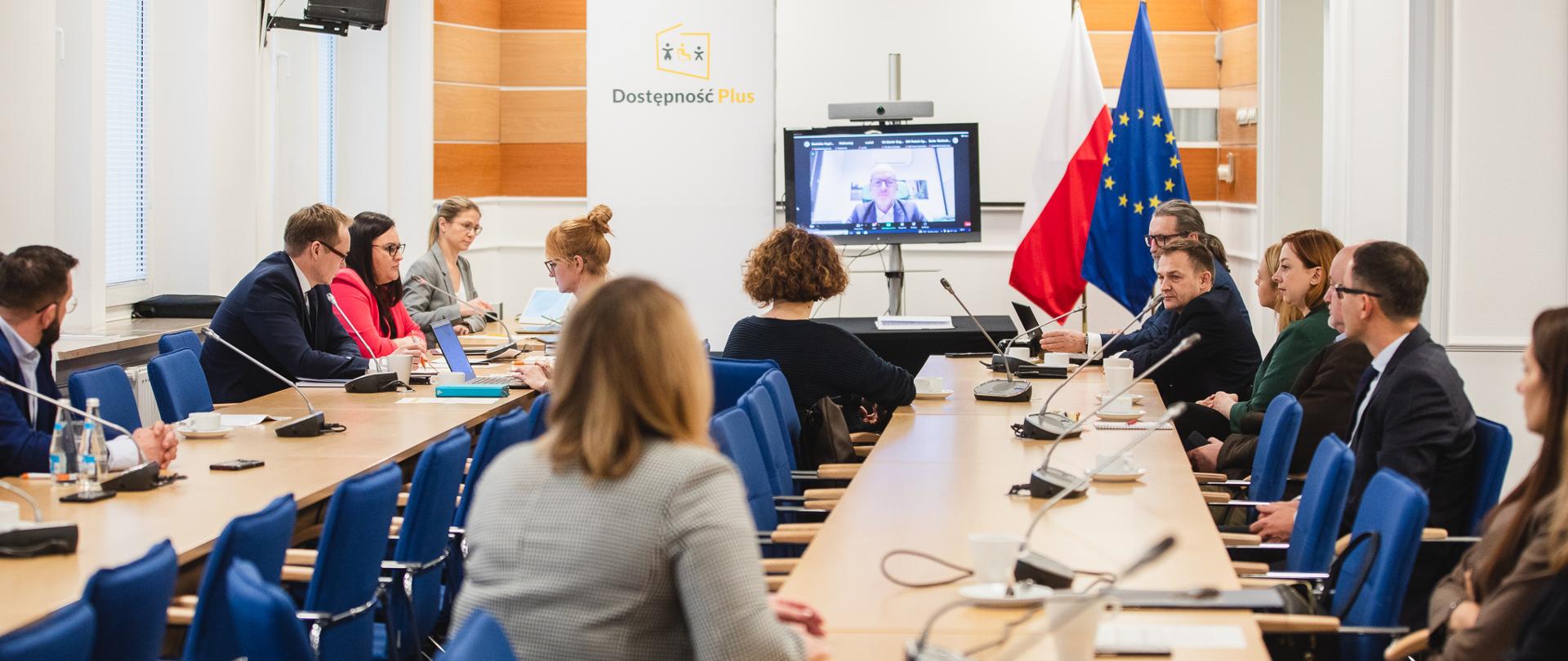 W sali przy stołach siedzą osoby. Przy ścianie monitor, flagi PL i UE i ścianka reklamowa z nadrukiem "Dostępność Plus".