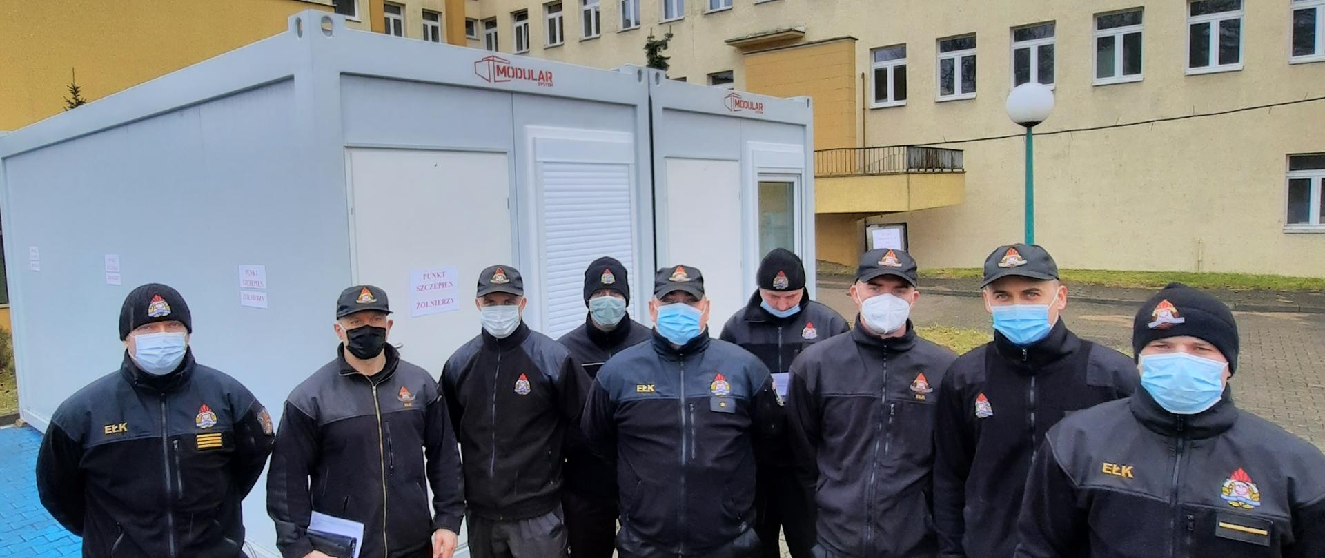 Zdjęcie przedstawia strażaków pozujących na tle punktu szczepień zorganizowanego na bazie kontenerów mieszkalnych, w tle Szpital Wojskowy