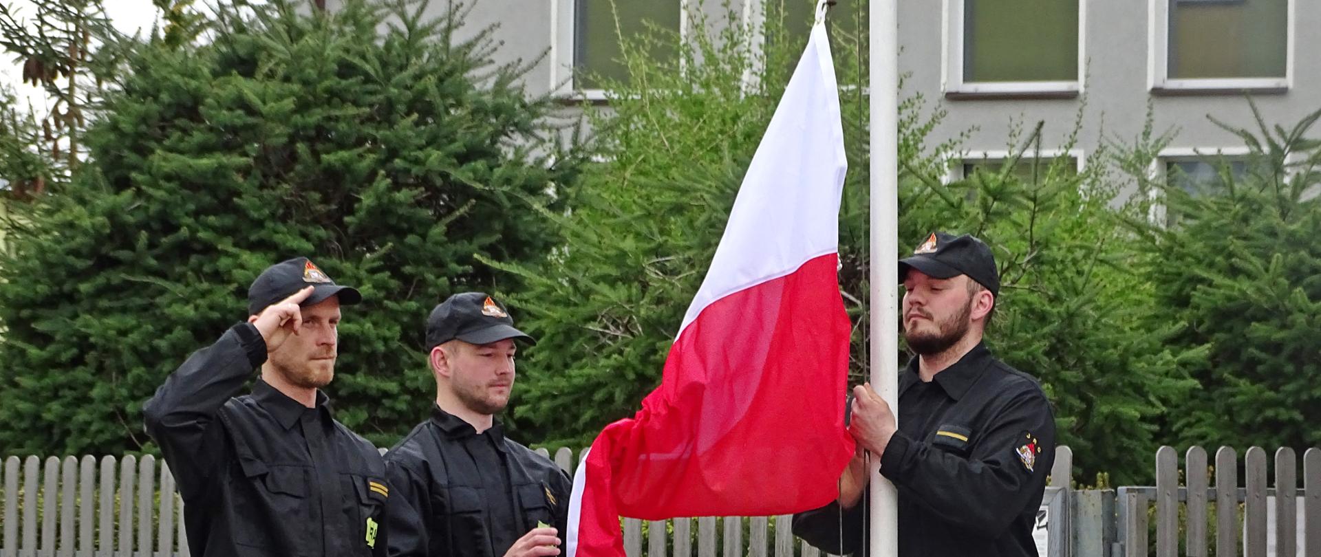 Zdjęcie wykonano w ciągu dnia przed budynkiem Komendy Powiatowej Państwowej Straży Pożarnej w Sępólnie Krajeńskim. Strażacy ubrani w mundury koszarowe stoją przed masztem na który wciągają flagę Rzeczypospolitej Polski.