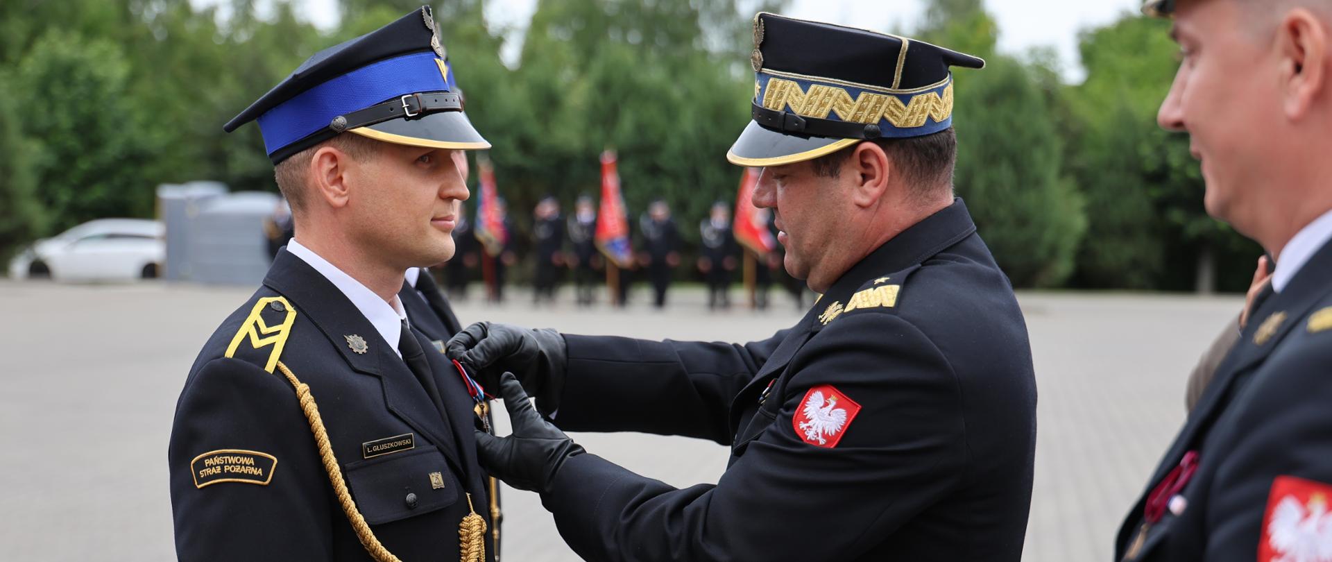 Moment dekorowania medalem podczas Powiatowych Obchodów Dnia Strażaka. Mazowiecki Komendant Wojewódzki nadbryg. Artur Gonera w czarnych rękawiczkach przypina odznaczenie do piersi stojącego na baczność młodszego oficera. Odznaczany strażak ma na naramiennikach dystynkcje kapitana. Po prawej stronie, na pierwszym planie, widoczny jest częściowy profil innego oficera trzymającego w dłoniach etui lub dokumenty do wyróżnień. W tle widać zarys placu komendy oraz poczty sztandarowe.