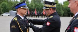 Moment dekorowania medalem podczas Powiatowych Obchodów Dnia Strażaka. Mazowiecki Komendant Wojewódzki nadbryg. Artur Gonera w czarnych rękawiczkach przypina odznaczenie do piersi stojącego na baczność młodszego oficera. Odznaczany strażak ma na naramiennikach dystynkcje kapitana. Po prawej stronie, na pierwszym planie, widoczny jest częściowy profil innego oficera trzymającego w dłoniach etui lub dokumenty do wyróżnień. W tle widać zarys placu komendy oraz poczty sztandarowe.