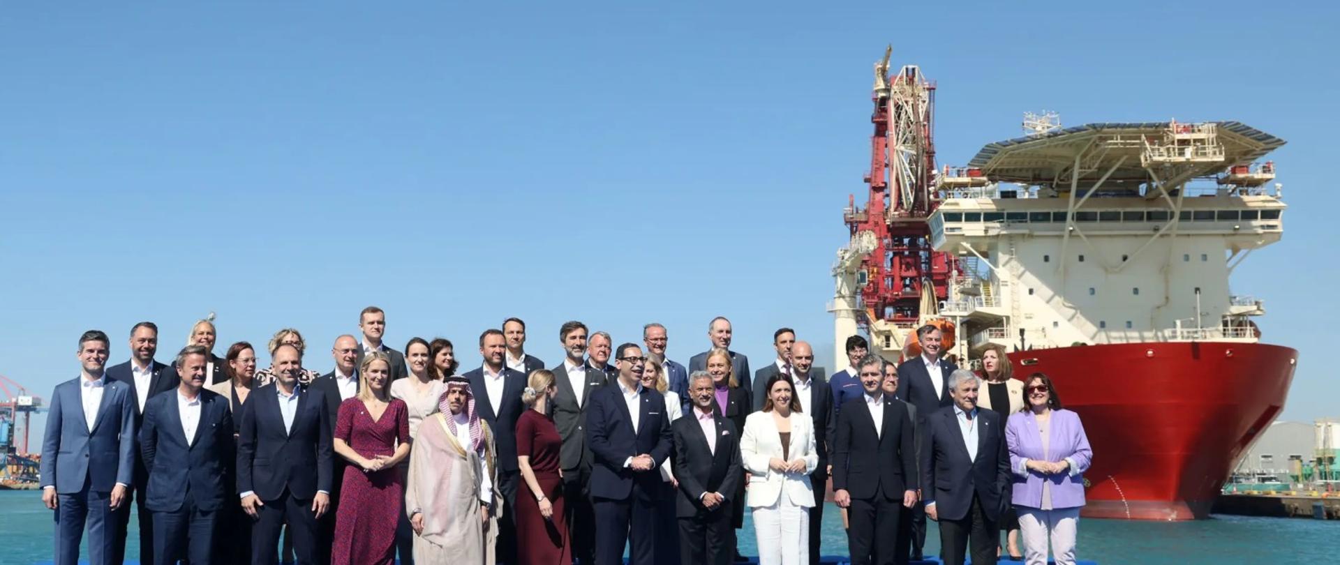 Participants of an informal meeting of foreign ministers pose for a family photo at a port. Most are dressed in suits and formal attire. A large red-and-white ship and a clear blue sky are visible in the background