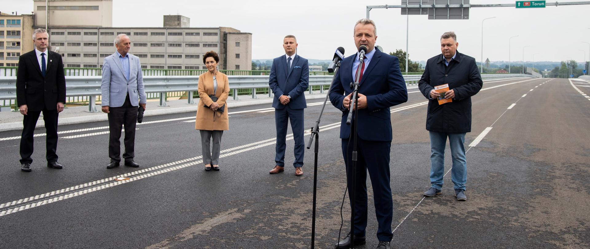 grupa osób stojąca na drodze, jeden mężczyzna przemawia przy mikrofonie
