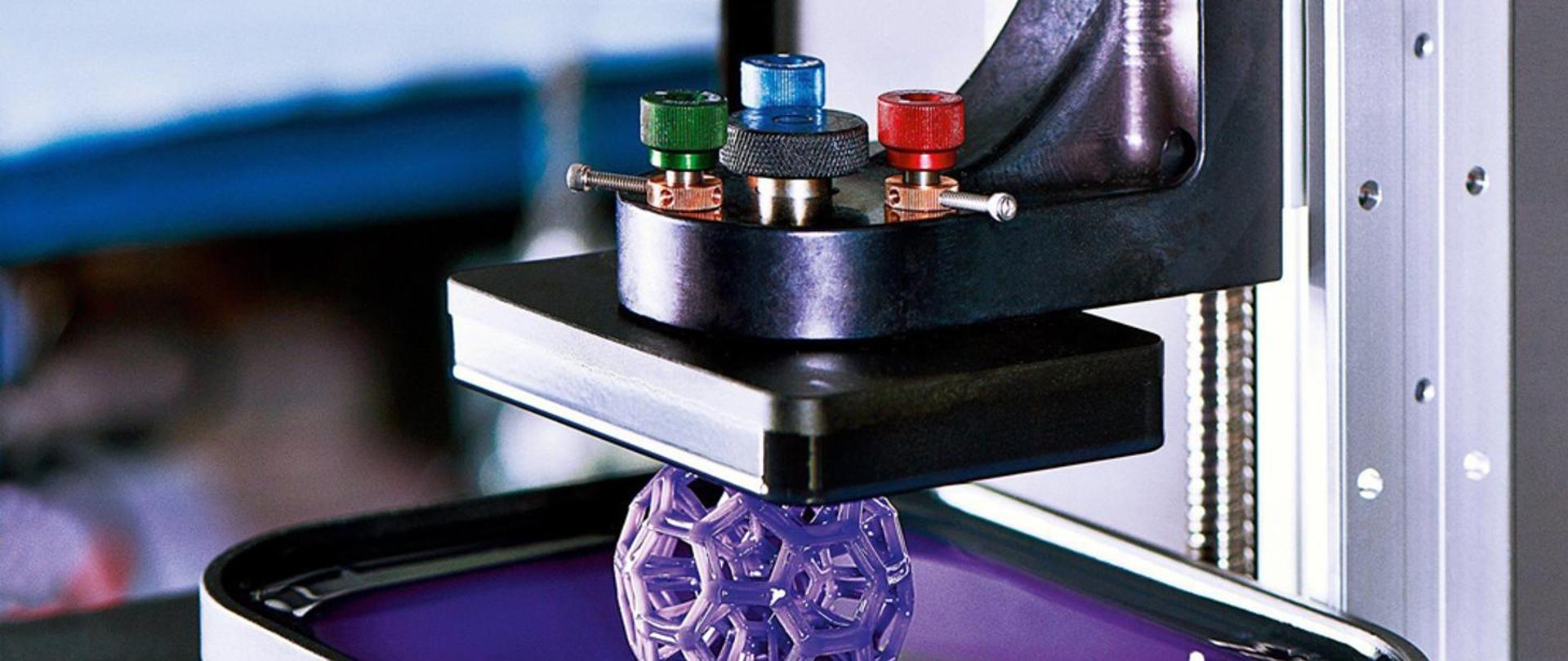 Na zdjęciu widoczna jest drukarka 3D w trakcie pracy, wykorzystująca technologię druku z żywicy światłoutwardzalnej. Urządzenie podnosi z platformy roboczej fioletowy, przestrzenny model o skomplikowanej, ażurowej strukturze. W tle można zauważyć elementy konstrukcyjne maszyny oraz kolorowe pokrętła regulacyjne.