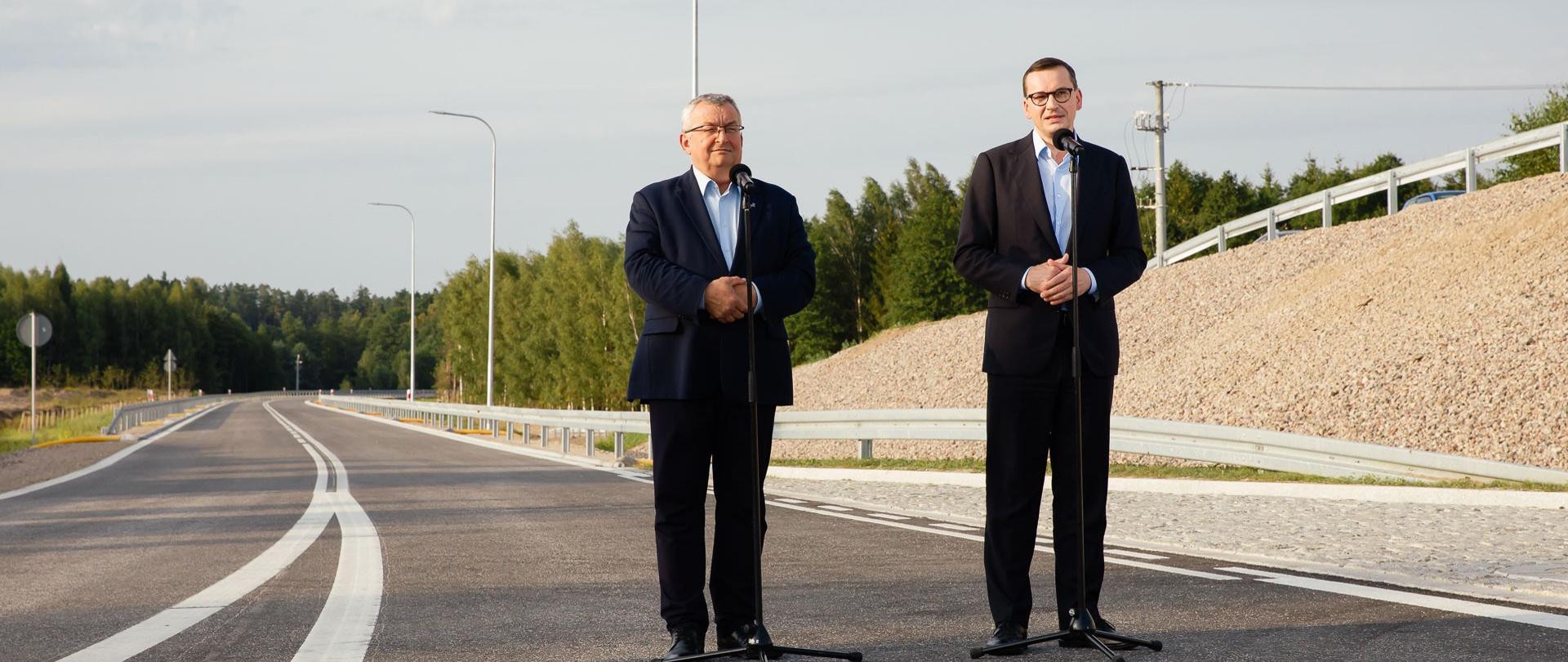 Premier oraz minister Andrzej Adamczyk stoją podczas konferencji prasowej na tle obwodnicy.