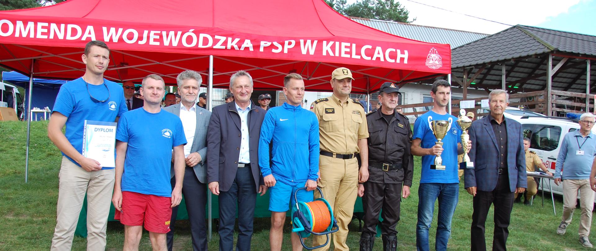 a pierwszym planie zdjęcia widać 9 osób stojących w szeregu na wzniesieniu pokrytym trawnikiem. Są to mężczyźni wśród których znajdują się uczestnicy biorący udział w zawodach ubrani w niebieskie bluzy i koszulki oraz włodarze, szefowie służb mundurowych zarazem będący organizatorami przedsięwzięcia na zbiorniku wodnym w Chańczy. Osoby w niebieskich strojach to drużyna zwycięzców zawodów nagrodzona pucharami, dyplomem i liną asekuracyjną na bębnie.
Za plecami wymienionych osób znajduje się czerwony namiot z napisem „KOMENDA WOJEWÓDZKA PSP W KIELCACH”