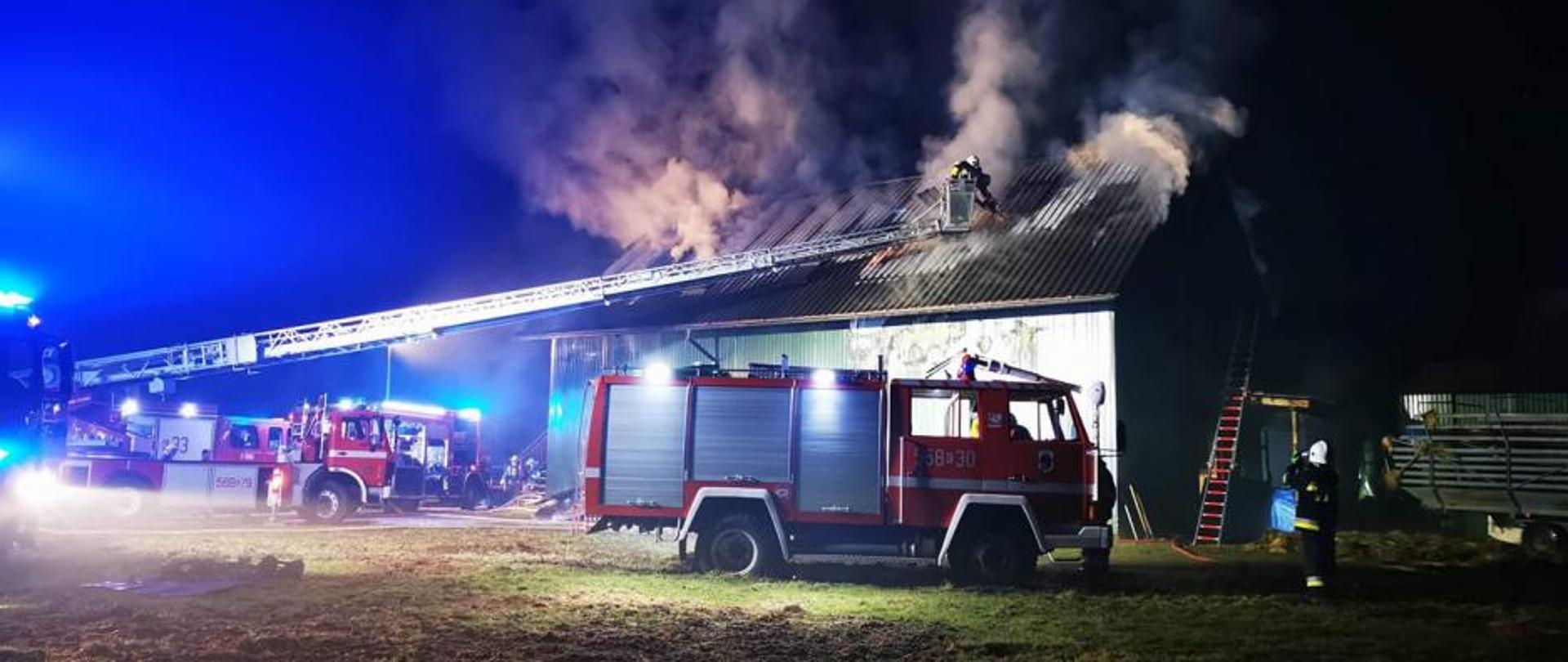 Na zdjęciu budynek jednokondygnacyjny z poddaszem objęty pożarem. Widoczny wydobywający się dym z dachu. Na zdjęciu również rozstawiona drabina pożarnicza oraz samochody ratowniczo - gaśnicze w czasie prowadzonych działań