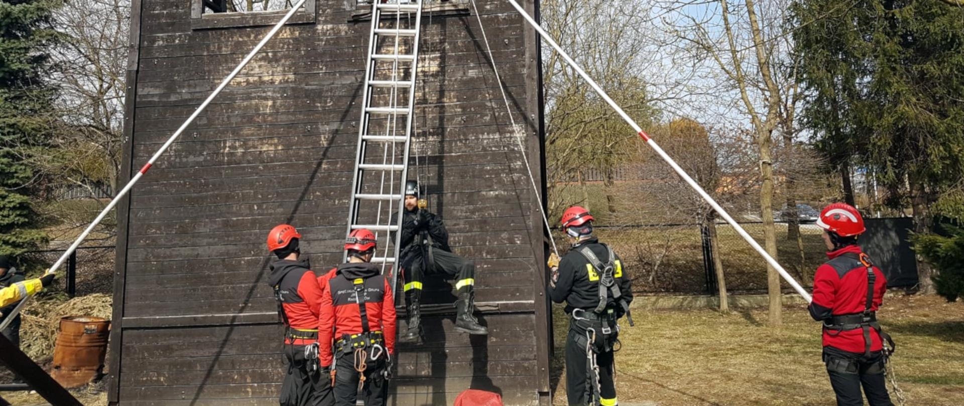 Na zdjęciu widoczna jest ściana wspinalni, na której ćwiczy pięciu strażaków ze specjalistycznej grupy ratownictwa wysokościowego. Wszyscy ubrani są w szelki zabezpieczające i kaski ochronne. Jeden ze strażaków zwieszony jest na linie przy ścianie, obok dostawiona jest drabina przystawna.
