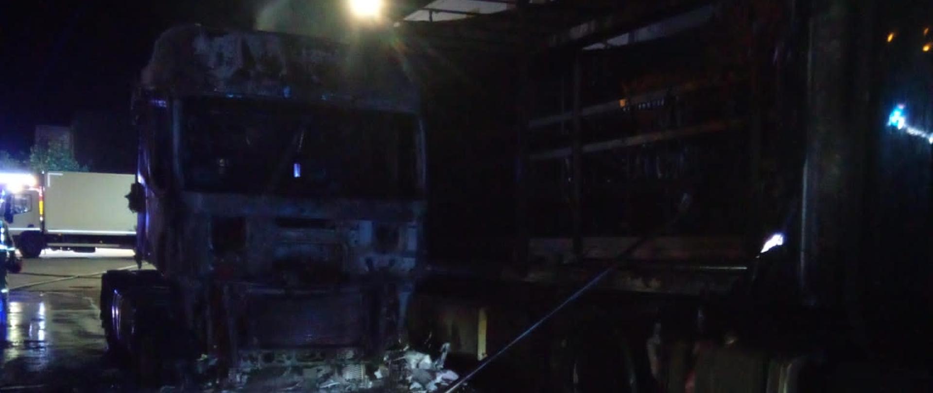 Zdjęcie przedstawia dwa spalone ciągniki siodłowe z naczepami. Po lewej widać spaloną naczepę a w głębi przód drugiego zniszczonego przez ogień pojazdu.