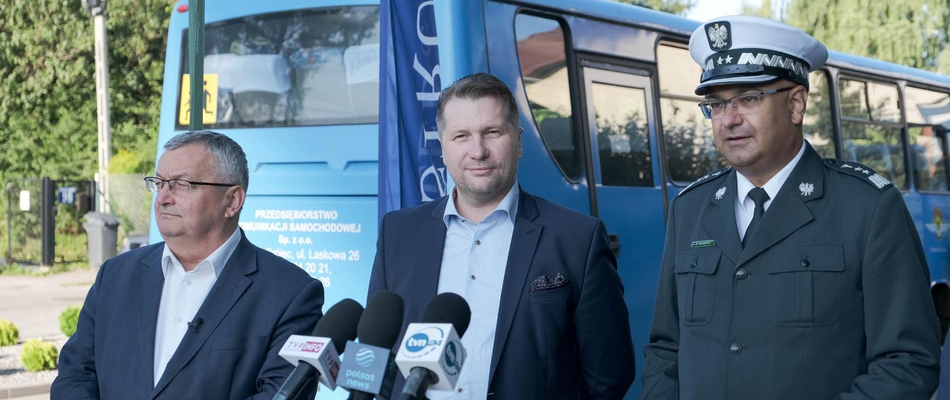 Minister Edukacji i Nauki Przemysław Czarnek wraz z Inspektorem Transportu Drogowego i Ministrem Infrastruktury stoją przed mikrofonem podczas briefingu prasowego dotyczącego bezpiecznego powrotu do szkół. W tle niebieski autokar