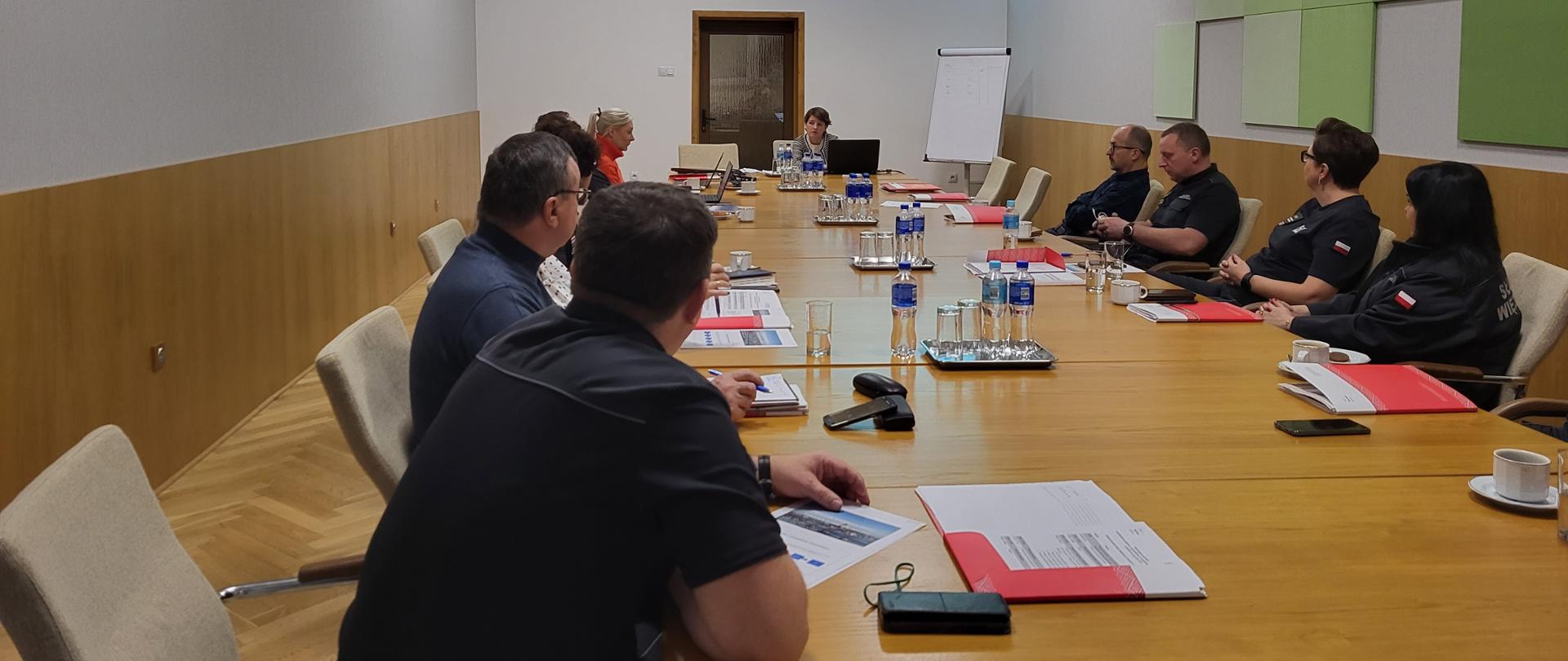 Długi brązowy stół konferencyjny. Po jego bokach przedstawiciele Operatora programu oraz Służby Więziennej na szarych krzesłach. Na ścianie przedniej wyświetlona prezentacja. Pod prezentacją biała tablica z flipchartami. Na lewej ścianie zegar. Na prawej ścianie zielona mozaika.