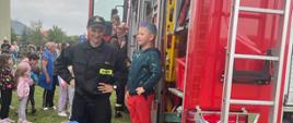 Zgromadzone wokół samochodu gaśniczego dzieci. Jeden z chłopców stoi na wysuniętym podeście w asyście strażaka. 