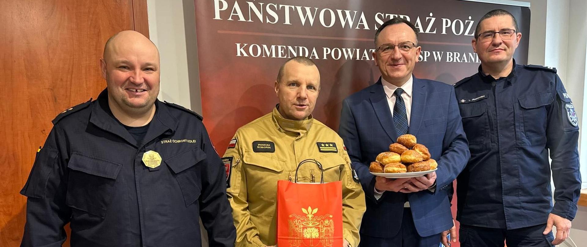 W gabinecie komendanta powiatowego spotkali się przedstawiciele służb oraz burmistrz Braniewa. Burmistrz trzyma talerz pączków.