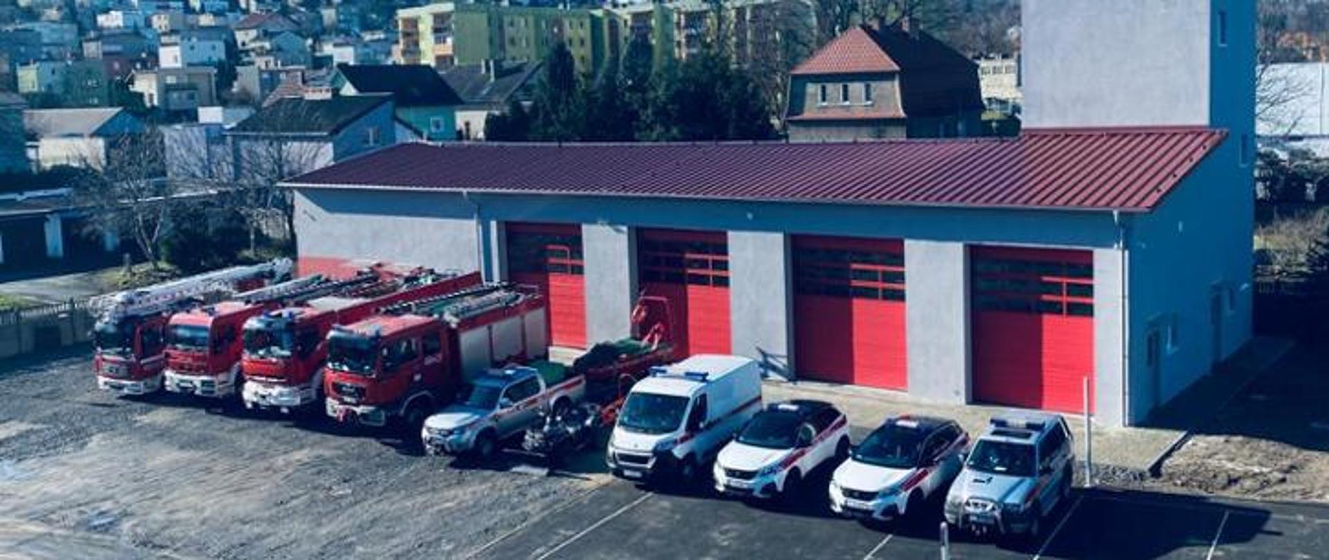 Widok z góry na budynek strażnicy Państwowej Straży Pożarnej z placem manewrowym i zaparkowanymi pojazdami służbowymi. Przed czterema czerwonymi bramami garażowymi stoją samochody ratowniczo-gaśnicze, pojazdy specjalistyczne oraz samochody wykorzystywane do realizacji zadań służbowych.