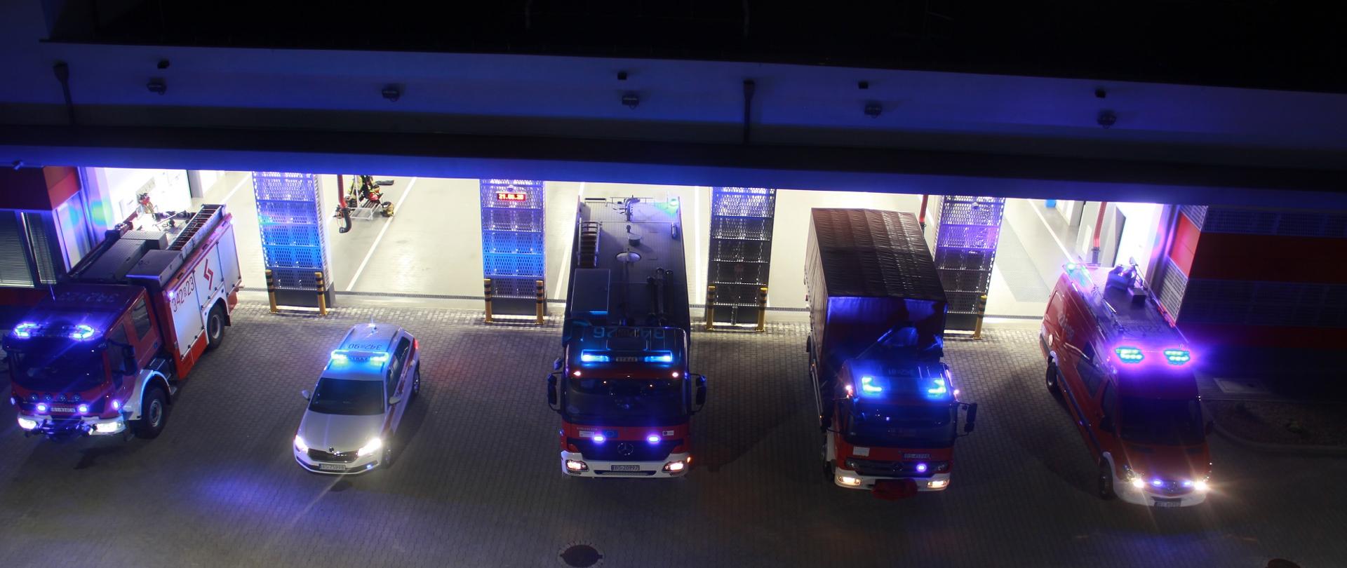 Na zdjęciu widać w nocy przed bramami garaży 5 samochodów strażackich z włączonymi sygnałami świetlnymi.