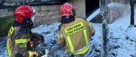 Dwóch strażaków w strojach bojowych gasi pożar zniszczonego budynku na stropie dachu