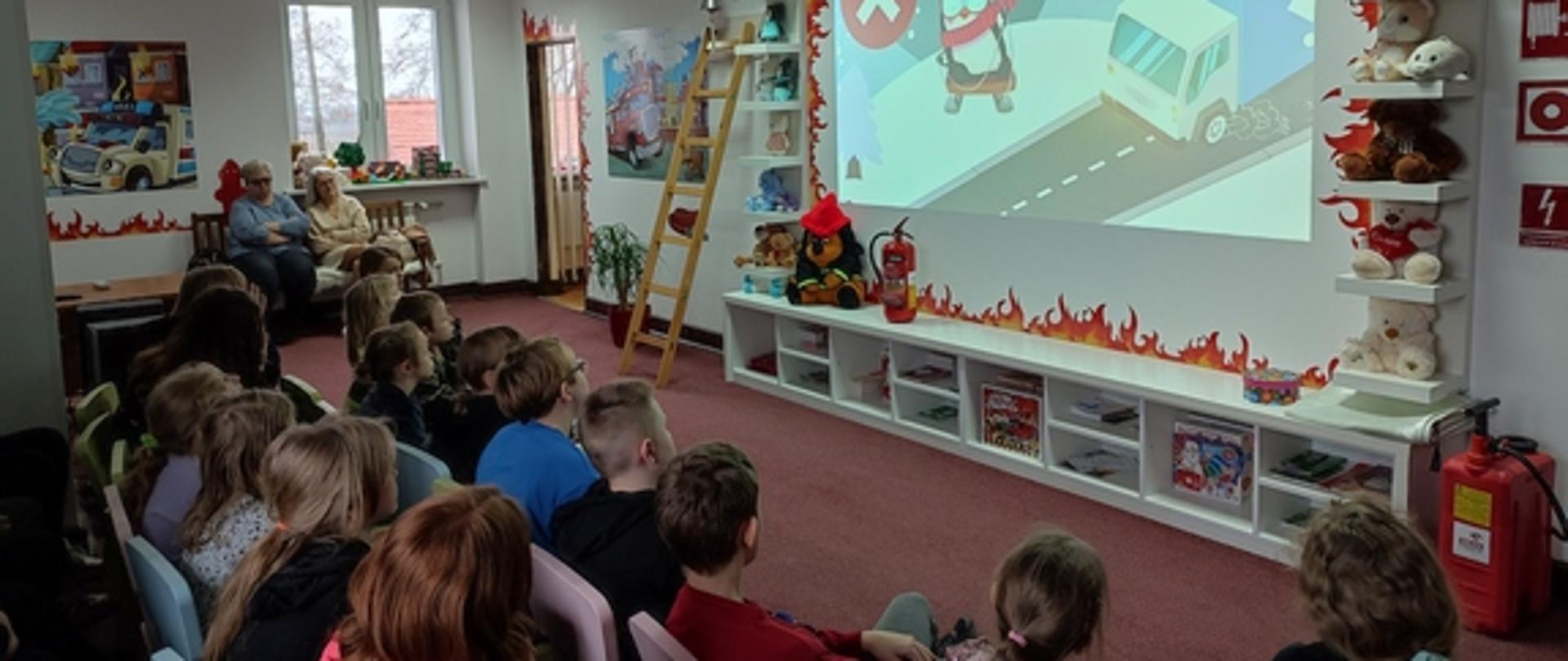 Dzieci ze Szkoły Podstawowej nr 3 w Łowiczu siedzą na krzesełkach i oglądają film wyświetlany na ścianie w tle drabina drewniana oraz okno pod którym siedzą dwie opiekunki na ścianach kolorowe infografiki na pierwszym planie czerwona hydronetka na podłodze ciemnobrązowa wykładzina 