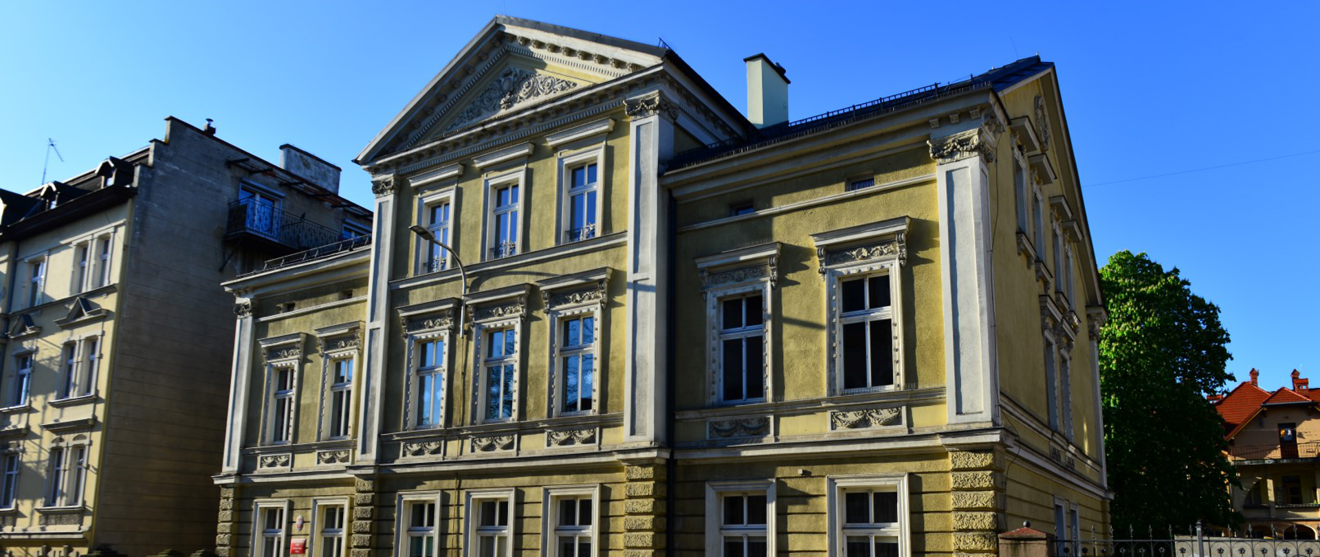 Zabytkowy budynek Państwowej Szkoły Muzycznej I stopnia w Kłodzku, z bogato zdobioną fasadą, wysokimi oknami i trójkątnym frontonem. Przed budynkiem znajduje się chodnik i żółte barierki, a w tle drzewa oraz zabudowa miejska. Zdjęcie wykonano w słoneczny dzień.