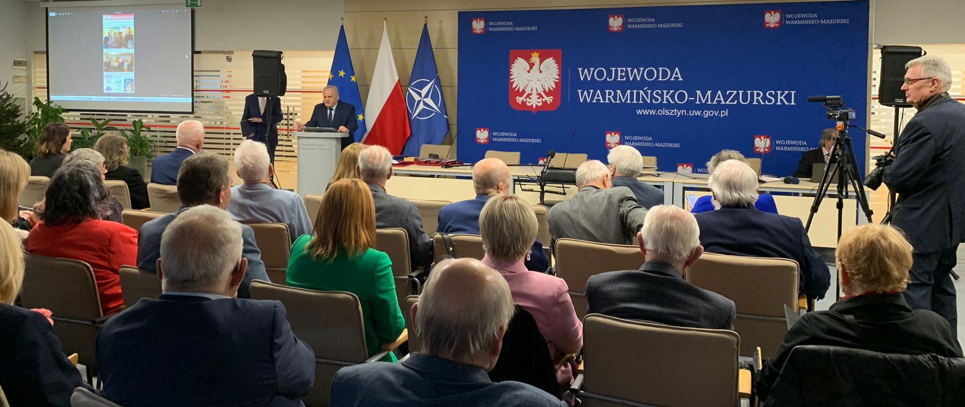 Zdjęcie przedstawia kilkanaście osób siedzących na krzesłach w sali, w której przemawia jeden mężczyzna 