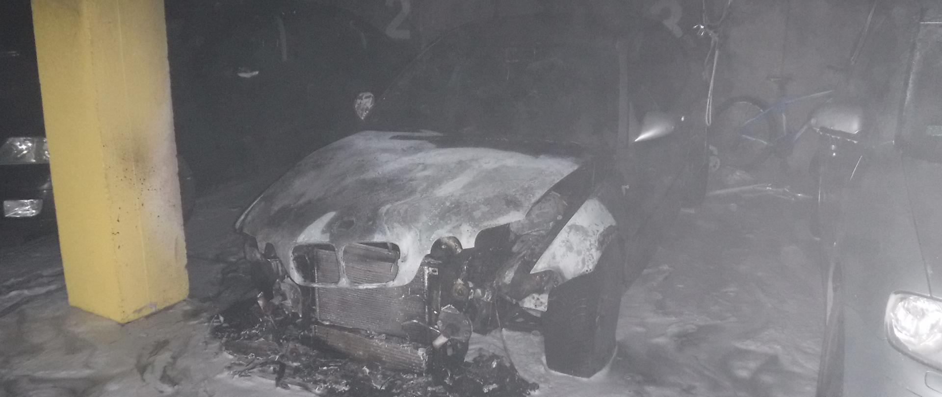 Zdjęcie przedstawia garaż podziemny w budynku wielorodzinnym. Na zdjęciu widać spalony pojazd osobowy oraz cześć stojące obok niego dwóch innych pojazdów osobowych. Na zdjęciu widać że doszło do spalenia całej komory silnika, pojazd ma stopione światła przednie i cały pas przedni. Za pojazdem widać rower. Na posadzce widać piane gaśniczą wykorzystana przez strażaków do gaszenia palącego się pojazdu.