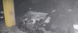 Zdjęcie przedstawia garaż podziemny w budynku wielorodzinnym. Na zdjęciu widać spalony pojazd osobowy oraz cześć stojące obok niego dwóch innych pojazdów osobowych. Na zdjęciu widać że doszło do spalenia całej komory silnika, pojazd ma stopione światła przednie i cały pas przedni. Za pojazdem widać rower. Na posadzce widać piane gaśniczą wykorzystana przez strażaków do gaszenia palącego się pojazdu.