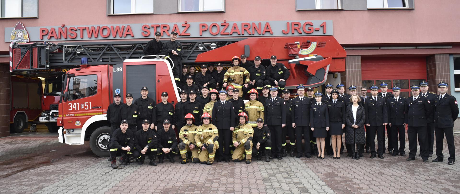 Na zdjęciu widać funkcjonariuszy PSP w umundurowaniu koszarowym, specjalnym oraz wyjściowym. Zdjęcie przed garażem JRG. Autodrabina.. Oświetlenie naturalne. Podłoże z kostki brukowej.