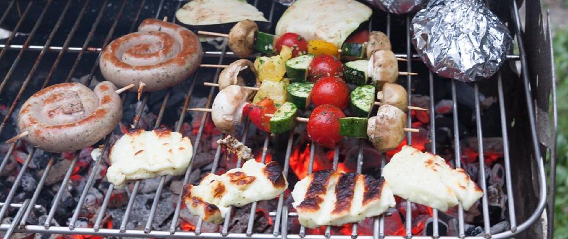 Grill z rozpalonym brykietem, na nim ruszt z grillowanymi potrawami: szaszłyki, ser, kiełbasa, cukinia, ziemniaki zawinięte w folię aluminiową.
