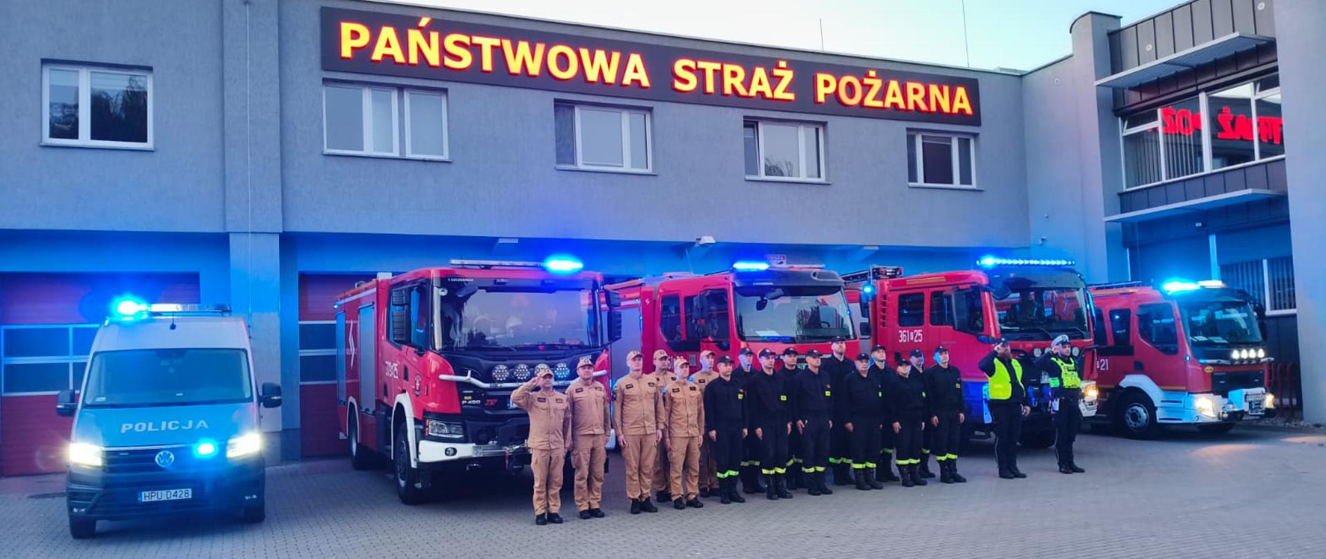 Strażacy stoją na placu przed remizą i minutą ciszy oddali hołd zmarłym strażakom. Z tyłu widać samochód strażacki na włączonych sygnałach świetlnych i dźwiękowych