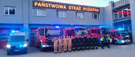 Strażacy stoją na placu przed remizą i minutą ciszy oddali hołd zmarłym strażakom. Z tyłu widać samochód strażacki na włączonych sygnałach świetlnych i dźwiękowych