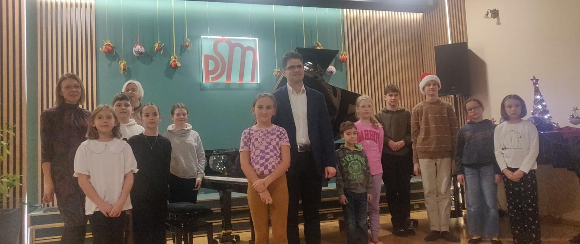 Na zdjęciu dzieci które uczestniczyły w warsztatach oraz nauczyciele i pan prowadzący w tle widoczne 2 fortepiany oraz ściana z wiszącymi ozdobami świątecznymi