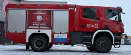 Czerwony wóz strażacki zaparkowany na zaśnieżonym placu przed budynkiem. Na boku pojazdu, na czerwonym tle, znajduje się numer "481M22". Pod numerem widnieje napis "KP PSP LIPSKO". Na lewej stronie wozu, na drzwiach schowka, widoczny jest napis "ZADBAJ O BEZPIECZEŃSTWO" oraz symbol czujnika dymu i czadu z napisem "ZAMONTUJ CZUJKĘ DYMU I CZADU". W tle, po lewej stronie, widoczny jest fragment budynku z elementami dachu pokrytego śniegiem i rynnami. Niebo jest pochmurne, ale jasne.