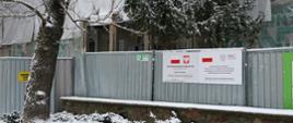 Mały fragment budynku w rusztowaniu przysłoniętym plandeką wykonawcy remontu. Widać zielone logo firmy GNKNIT. Przed budynkiem szarostalowy płot odgradzający plac budowy. Na nim dwie plansze umieszczone jedna obok drugiej, informujące o inwestycji i źródłach jej finansowania. Przed ogrodzeniem niski kamienny murek i drzewo. Na pierwszym planie krzewy obficie przysypane śniegiem. Za płotem zielona tuja, również pobielona śniegiem.