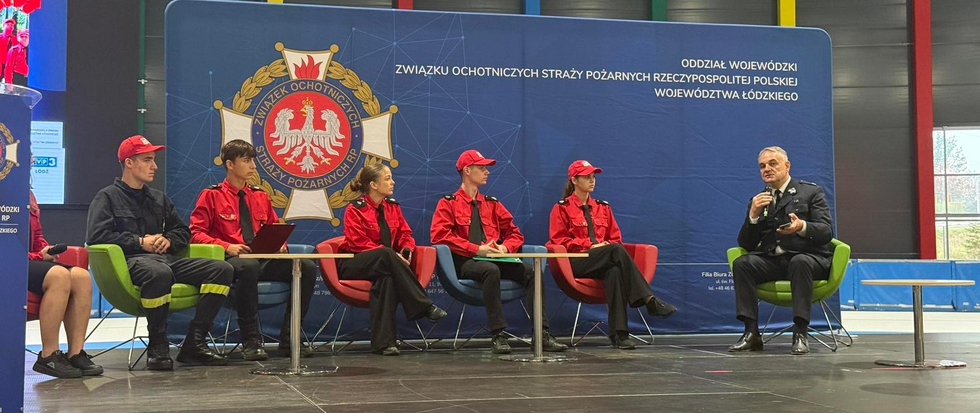 panel dyskusyjny z udziałem czterech młodych osób w czerwonych koszulkach z napisem "STRAŻ" i jednego mężczyzny w mundurze. Wszyscy siedzą na kolorowych fotelach na scenie przed wielkim banerem z logo Związku Ochotniczych Straży Pożarnych.