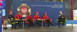 panel dyskusyjny z udziałem czterech młodych osób w czerwonych koszulkach z napisem "STRAŻ" i jednego mężczyzny w mundurze. Wszyscy siedzą na kolorowych fotelach na scenie przed wielkim banerem z logo Związku Ochotniczych Straży Pożarnych.