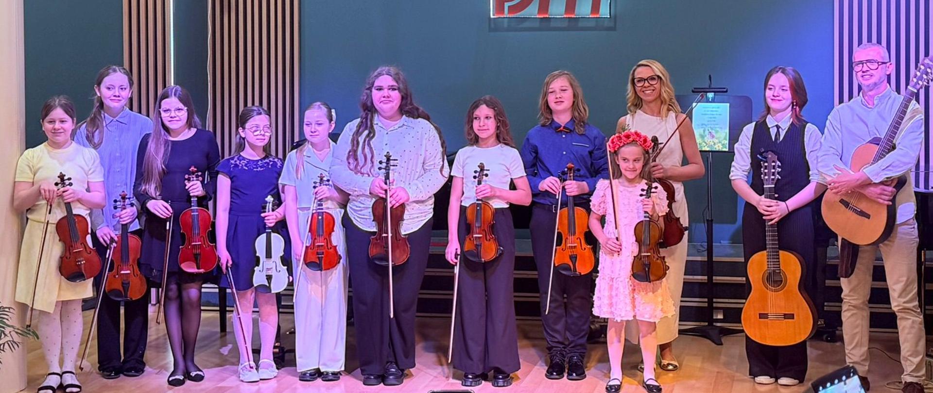 Zdjęcie uczniów ze skrzypcami w rękach oraz jedna uczennica trzyma gitarę a obok niej jej nauczyciel również trzyma gitarę w rękach wszyscy są uśmiechnięci i pozują do zdjęcia ana dole zdjęcia widać publiczność która robi zdjęcia swoimi telefonami