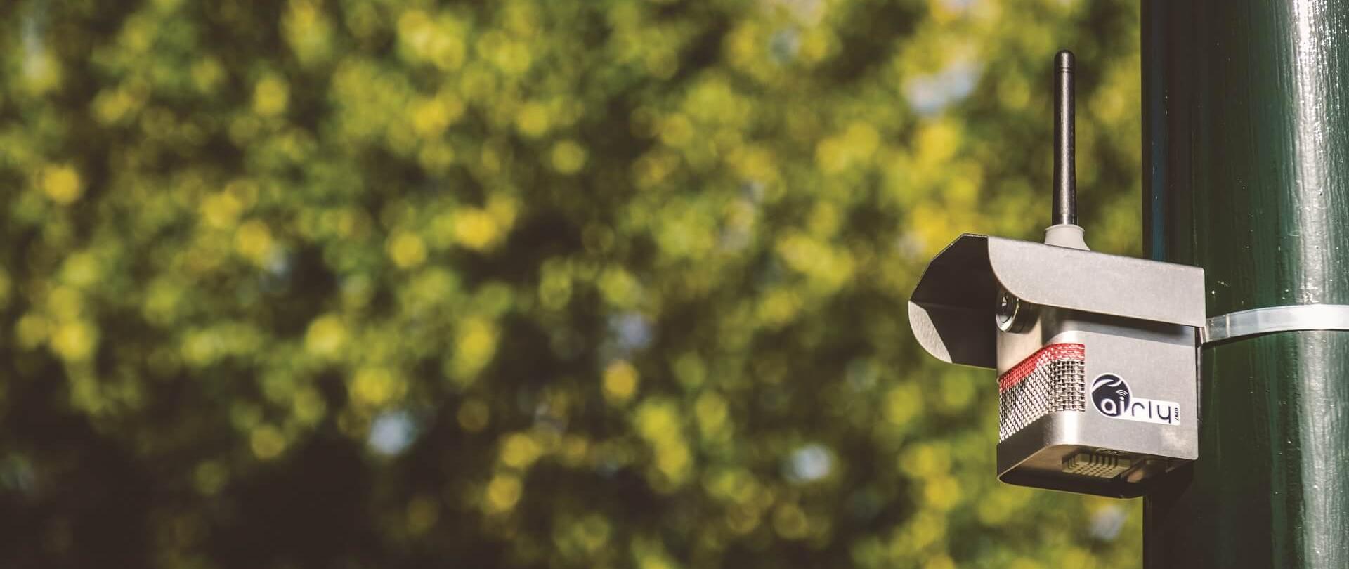 Czujnik AIRLY umieszczony na zielonym słupie, w metalowej obudowie, kształtem przypomina budkę dla szpaków z antenką