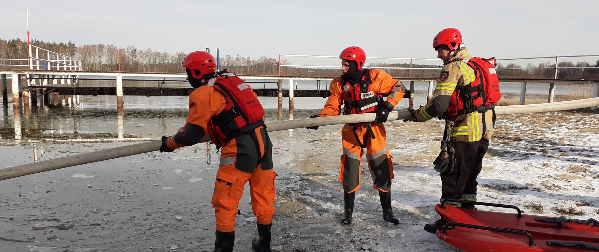 Zdjęcie przedstawia 3 strażaków w specjalnym ubraniu do pracy w wodzie, trzymają wąż, w tle pomost na akwenie wodnym, zamarznięta tafla lodu