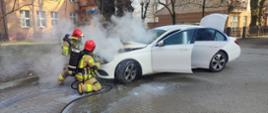 Na zdjęciu widać palący się biały samochód osobowy oraz dwóch strażaków podczas akcji gaśniczej. 