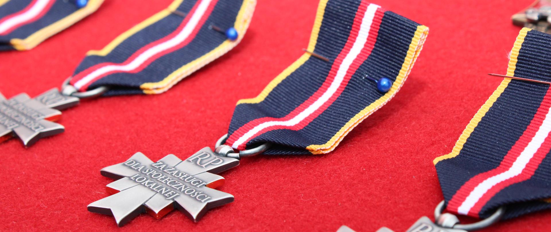 Zdjęcie przedstawia rzędy Odznak Świętego Floriana „Za Zasługi dla Społeczności Lokalnej”, ułożonych na czerwonym suknie. Srebrzyste krzyże z napisem „RP” zawieszone są na granatowo-żółto-czerwonych wstążkach i przygotowane do uroczystego wręczenia.