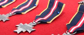 Zdjęcie przedstawia rzędy Odznak Świętego Floriana „Za Zasługi dla Społeczności Lokalnej”, ułożonych na czerwonym suknie. Srebrzyste krzyże z napisem „RP” zawieszone są na granatowo-żółto-czerwonych wstążkach i przygotowane do uroczystego wręczenia.
