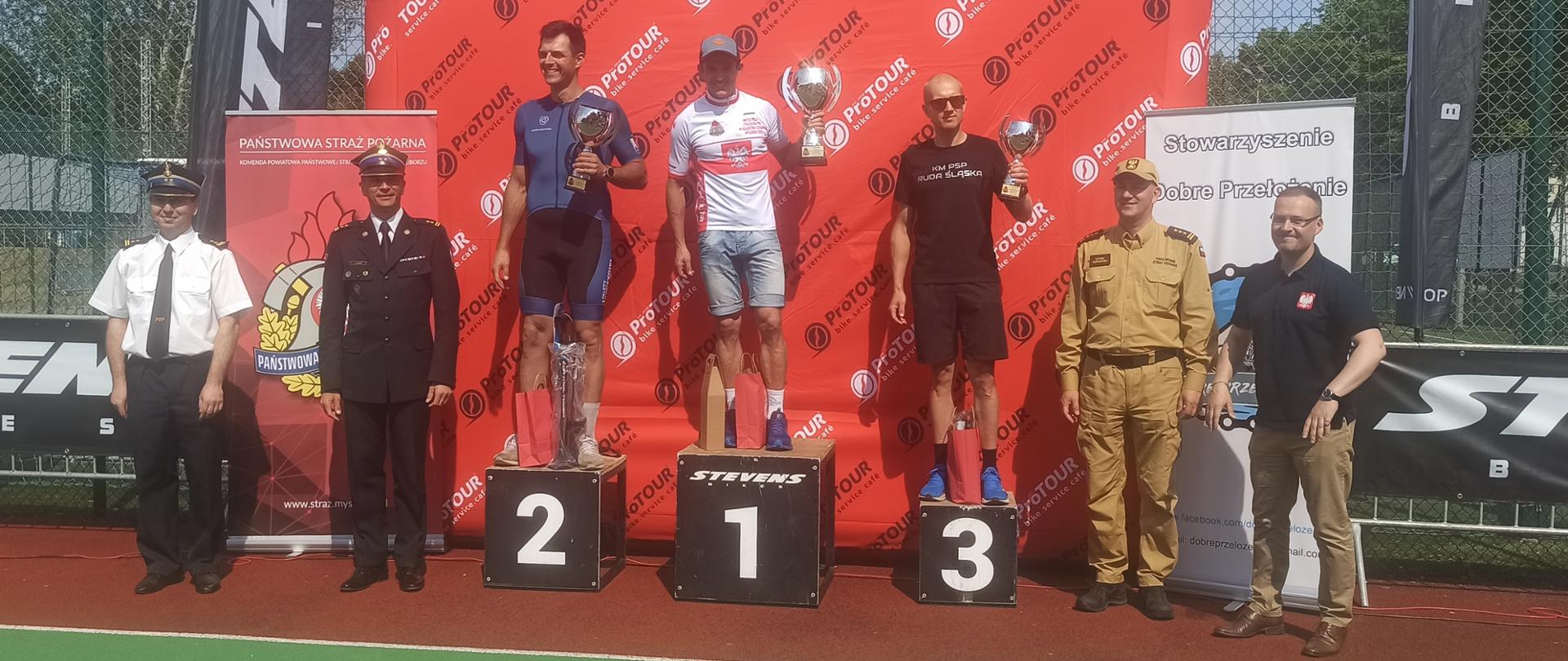 Zdjęcie przedstawia stojących na podium kolarzy z PSP. Na podium nr 1 stoi kolarz ubrany w białą koszulkę mistrza Polski i trzyma w lewej ręce puchar. Na podium drugim stoi kolarz ubrany w niebieski strój kolarski i w lewej dłoni trzyma puchar. Na trzecim miejscu podium stoi strażak ubrany w czarna koszulkę i lewej dłoni trzyma puchar. Po lewej stronie stoją dwaj strażacy jeden ubrany w mundur wyjściowy PSP i czapkę a drugi w białej koszuli z krawatem PSP. Po prawej stronie podium stoi strażak ubrany w piaskowy ubranie dowódcze z czapka z daszkiem. Za podium stoi wielki baner z napisem Pro TOUR a obok nich po lewej stronie baner Państwowej Straży Pożarnej a po prawej baner Stowarzyszenia Dobre Przełożenie.