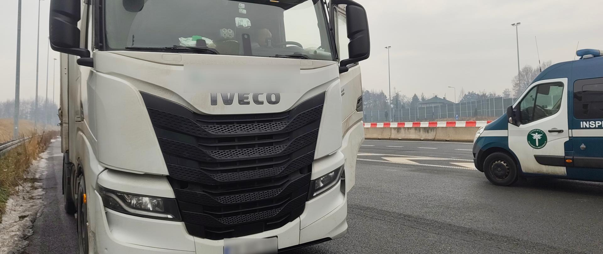 Na szerokim pasie drogowym lub parkingu przydrożnym widać zaparkowane wzdłuż pasów drogowych białe pojazdy ciężarowe z białymi naczepami. Obok nich zaparkowany jest również biało-zielony pojazd typu bus z oznaczeniami Inspekcji Transportu Drogowego. Najprawdopodobniej trwa kontrola drogowa. W tle widać ekrany akustyczne przy pasie drogowym oraz fragment lasu. Widoczne są ślady śniegu, niebo jest zachmurzone co sugeruje zimową aurę. 