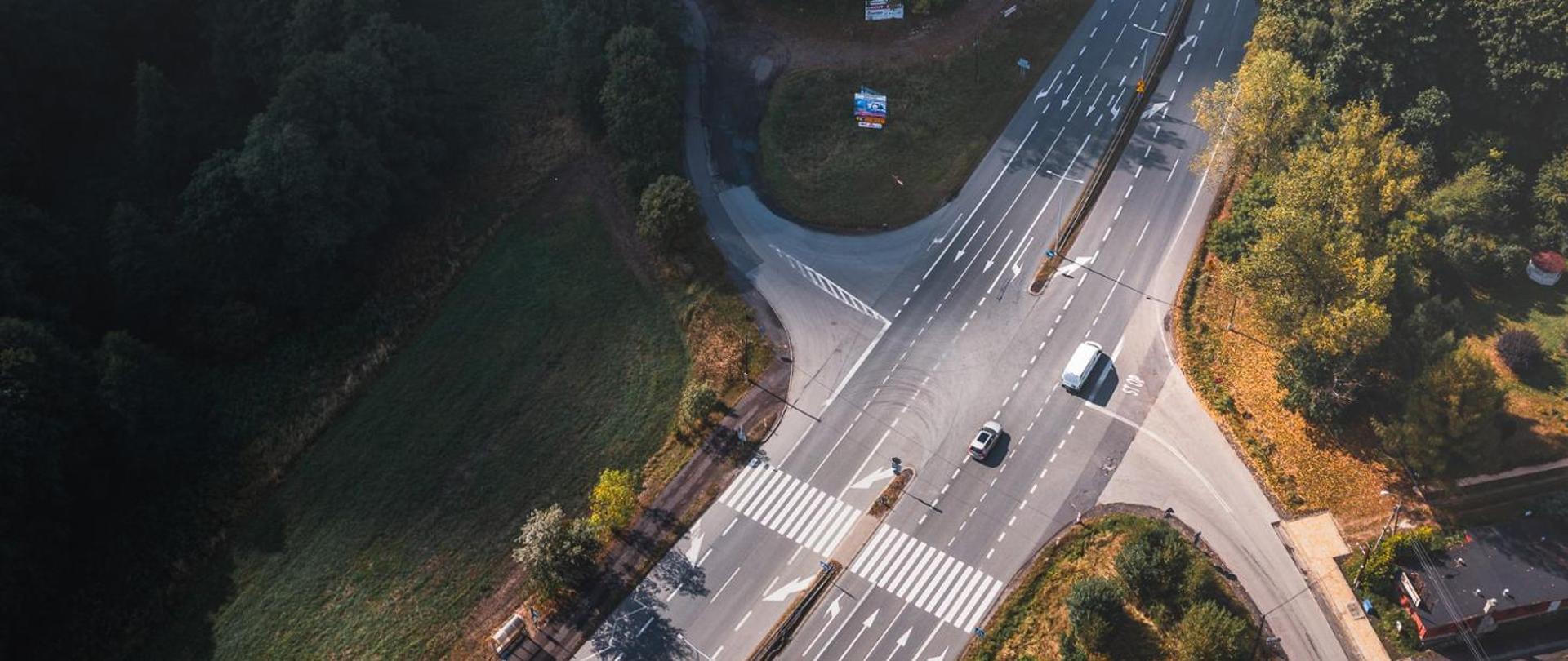 Zdjęcie lotnicze skrzyżowania na drodze DK81 otoczonego terenami zielonymi. Widoczne są wielopasmowe jezdnie, przejście dla pieszych oraz kilka pojazdów poruszających się w obu kierunkach. Wokół przebiegają ściany lasu i pojedyncze zabudowania.