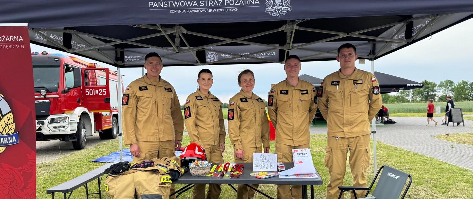 pod namiotem stoi pięciu strażaków w tym trzej mężczyźni i dwie kobiety przed nimi jest stolik na którym leży strój strażaka, cukierki, odblaski, kolorowanki i dyplomy W tle widać wóz strażacki