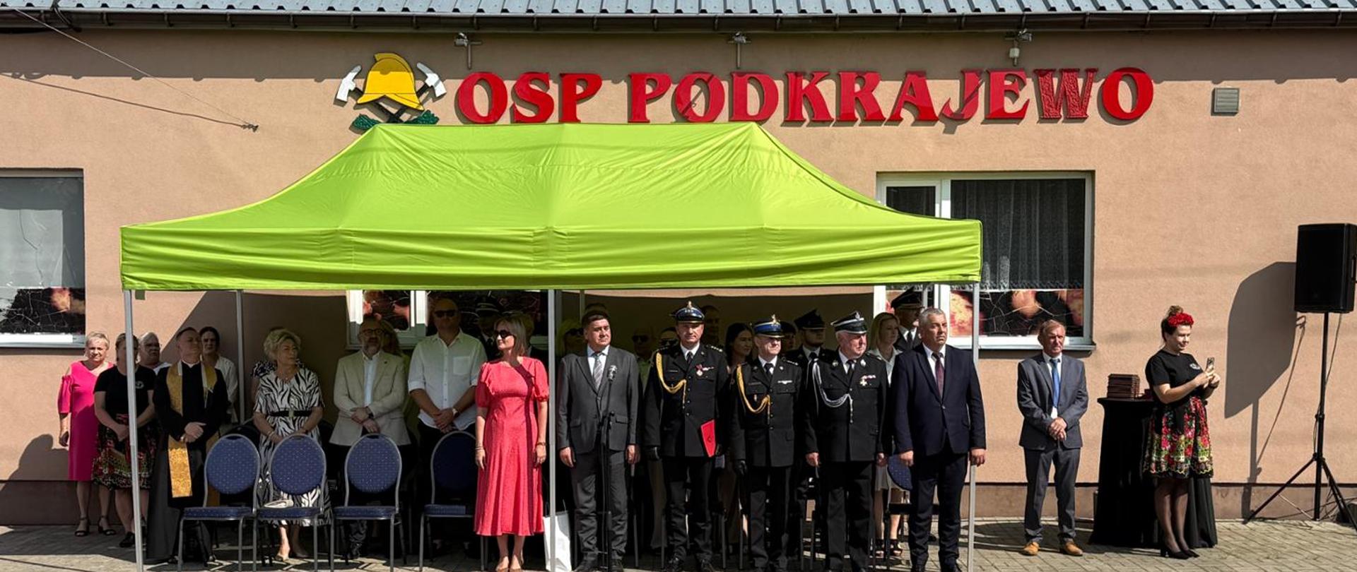 Uczestnicy uroczystości stoją pod jasno zielonym namiotem ustawionym przed budynkiem z napisem "OSP Podkrajewo" 