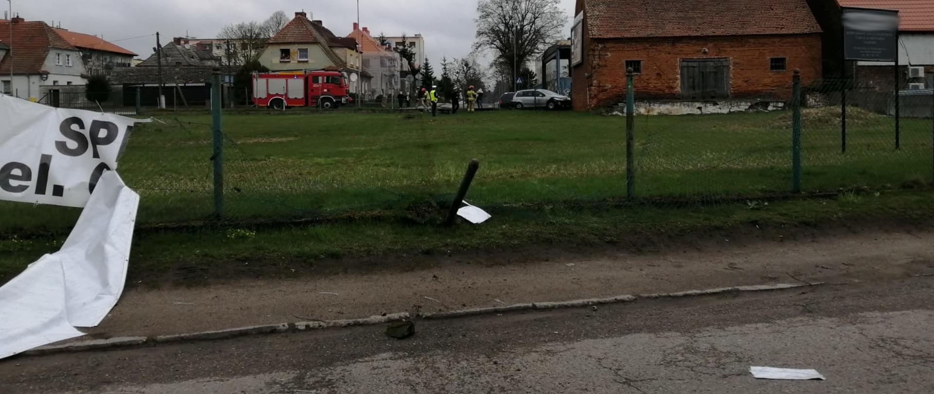 Fotografii przedstawia widok skrzyżowania ulicy Słowackiego i Wyzwolenia, ujęcie wykonane z ulicy cmentarnej. U dołu zdjęcia widoczny asfalt ulicy Cmentarnej, powyżej widać uszkodzone ogrodzenie z siatki i betonowych słupów, jeden słup jest mocno pochylony, siatka zerwana. Dalej za siatką jest zielony trawnik na którego końcu także znajduje się ogrodzenie za którym widoczne jest wymienione skrzyżowanie. Po prawej stronie skrzyżowania widoczny budynek z czerwonej cegły. Przy skrzyżowaniu widoczni są strażacy i policjanci oraz inne osoby. Skrzyżowanie oddalone jest o około 50 metrów od miejsca wykonania fotografii. Po lewej stronie skrzyżowania widoczny samochód gaśniczy. W tle zabudowania miasta oraz widoczny komin ciepłowni. Jest dzień, nieb jest zachmurzone.