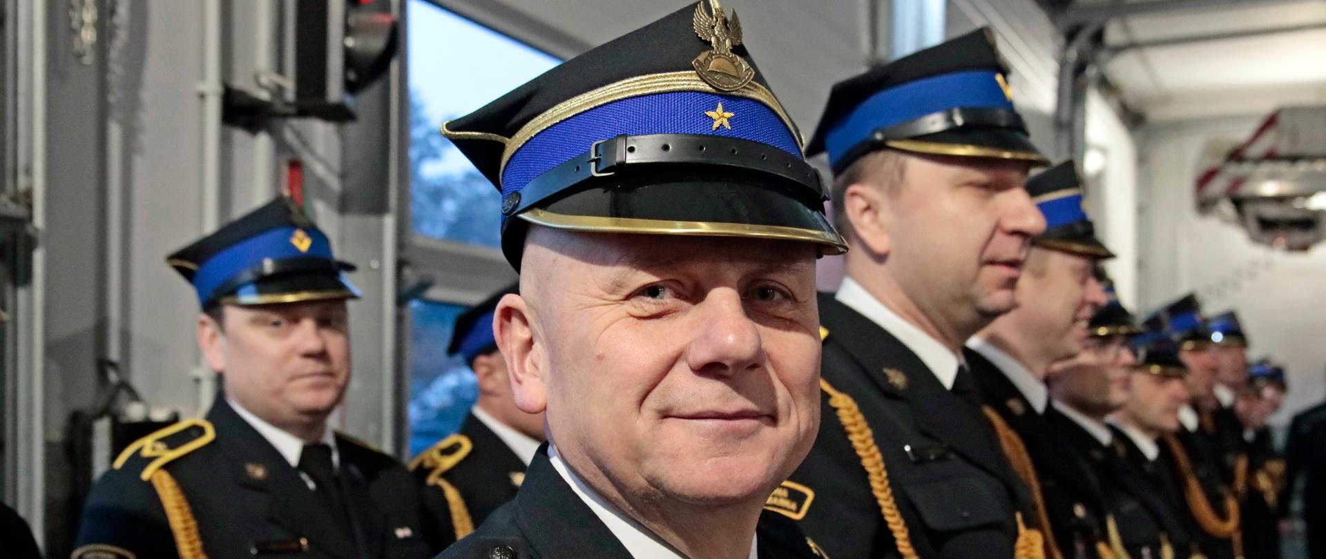 Strażacy w galowych mundurach i rogatywkach na głowie podczas zbiórki w pomieszczeniu