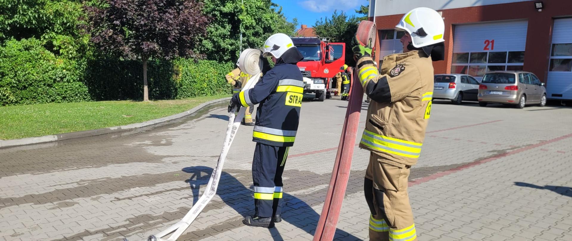 Dwóch strażaków zwija węże strażackie na placu brukowym w tle samochód pożarniczy i duży obiekt.