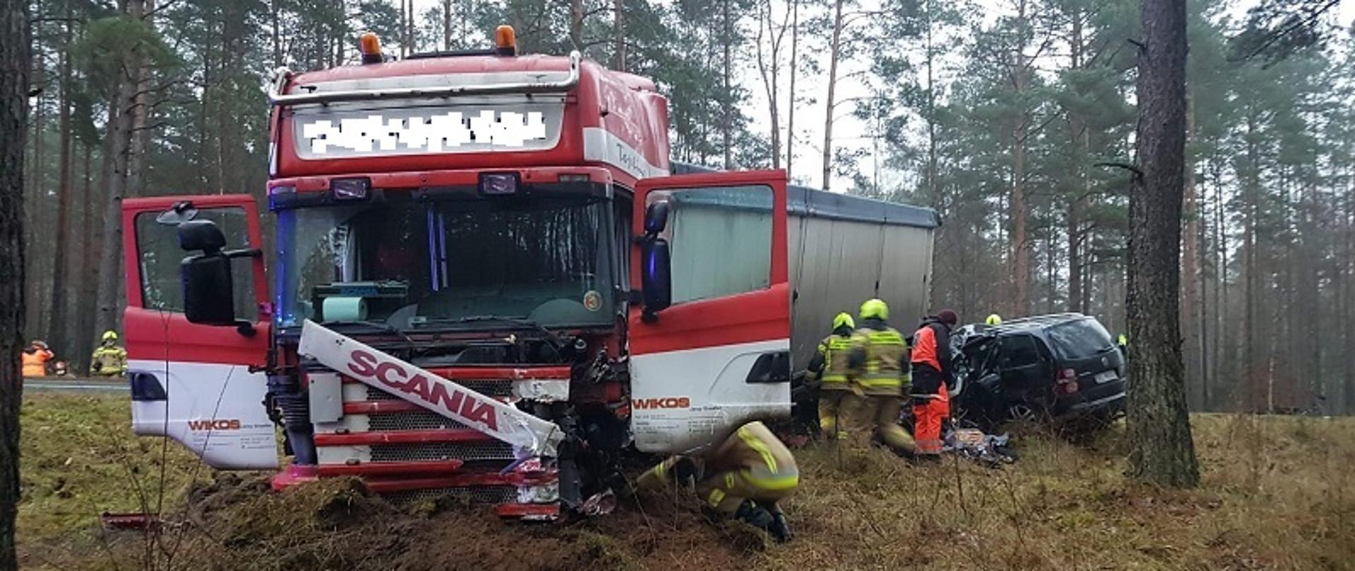 Zdjęcie przedstawiające samochód ciężarowy po zderzeniu z samochodem osobowym. Samochody częściowo po za pasem ruchu. Droga krajowa przebiegająca przez las. Strażacy wykonują czynności ratunkowe razem z ratownikami medycznymi.