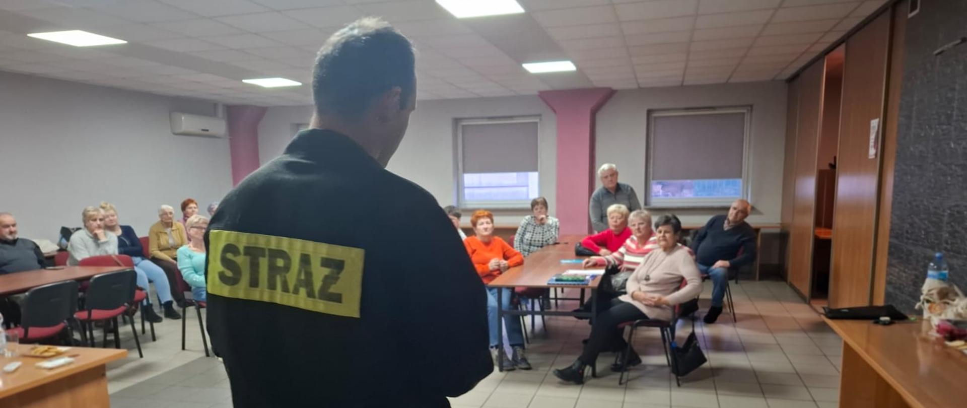 Strażak na sali wykładowej przed nim rozłożony fantom a w tle słuchający seniorzy